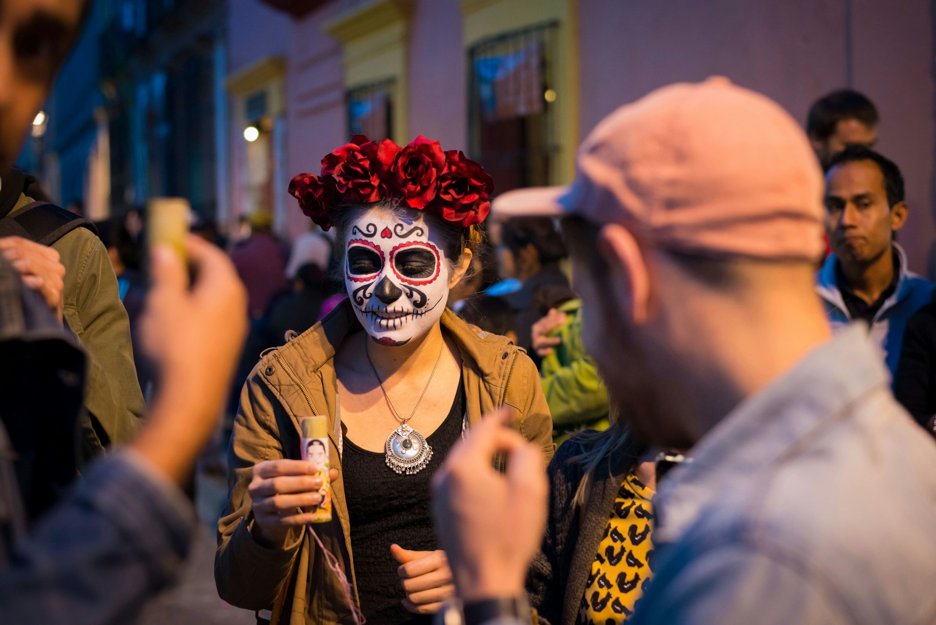 A young woman in a skeleton mask, holding an alcoholic drink while part of a crowd celebrating Day of the Dead in Oaxaca, Mexico.